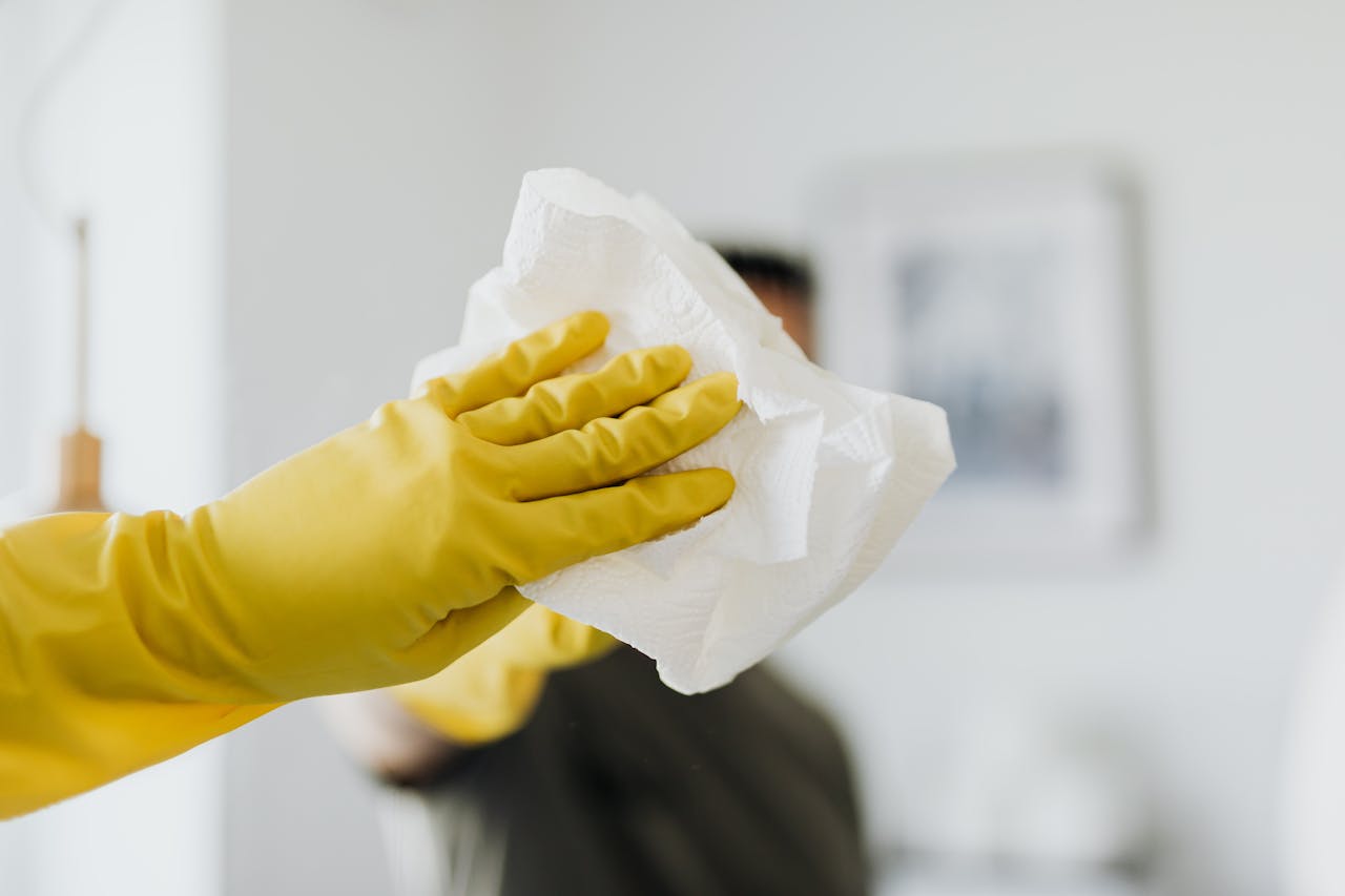 about-01 Close-up of a yellow-gloved hand cleaning a mirror with a cloth indoors.