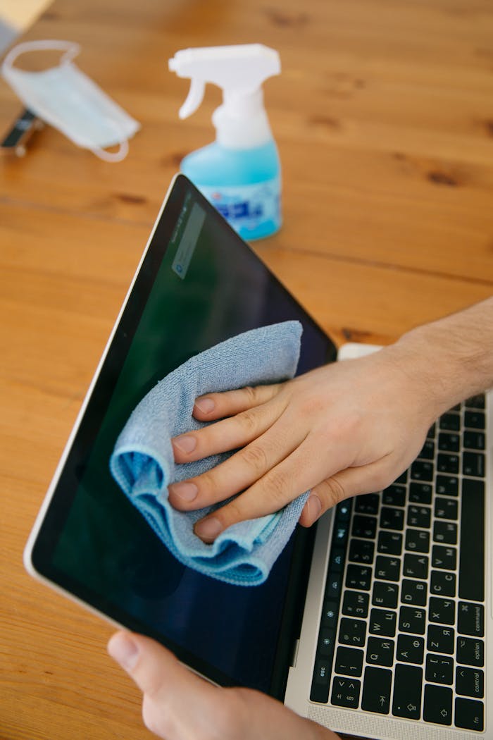 why-choose-us-03 A person wipes a laptop screen using a blue cloth and spray bottle on a wooden table for hygiene.