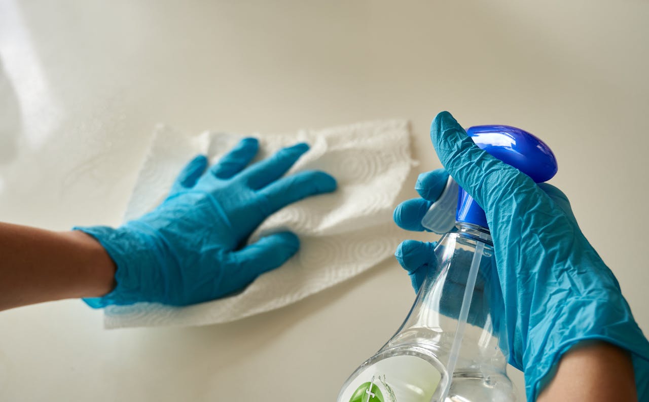 why-choose-us-02 Close-up of gloved hands using disinfectant spray and cloth for cleaning surface.
