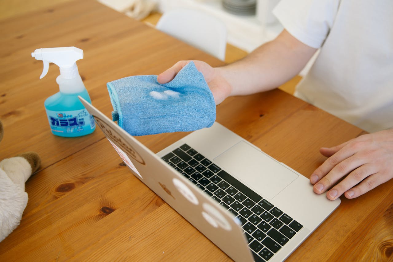 hero-02 Person disinfecting laptop with spray and cloth, ensuring cleanliness and hygiene indoors.